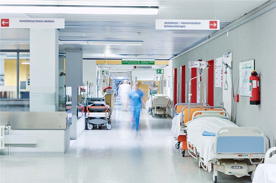 A hospital corridor with empty beds, medical equipment, and signs. A person in blue scrubs is walking down the hallway.