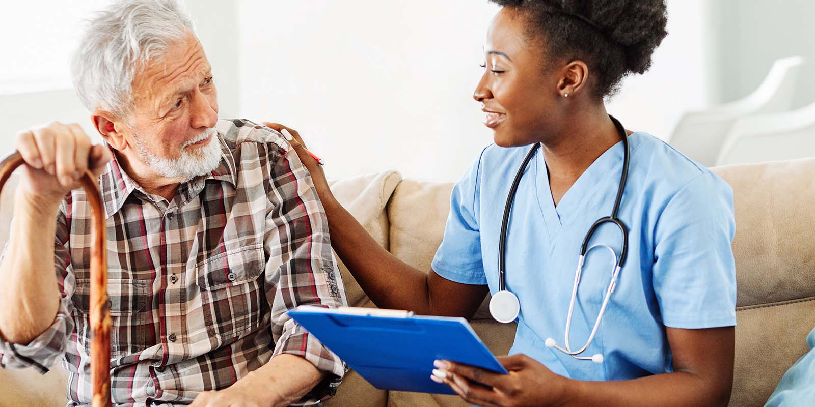 A nurse in blue scrubs holds a clipboard and talks to an elderly man with a cane, seated on a sofa.