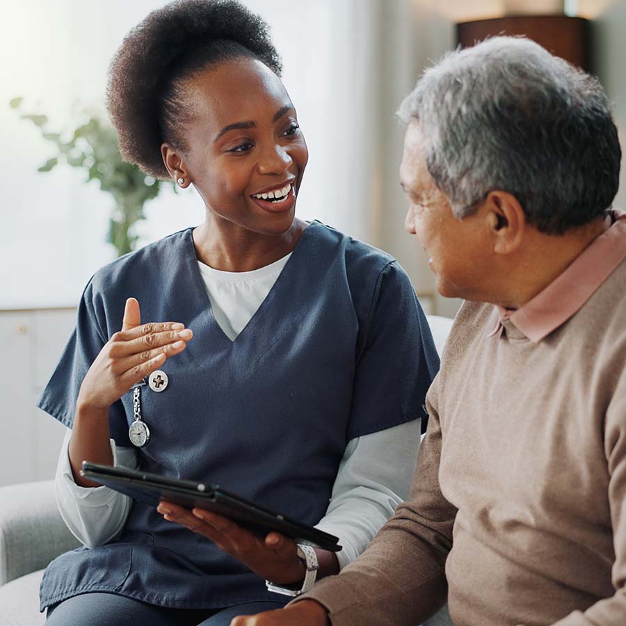 A healthcare professional talks with an elderly man, both seated indoors, holding a tablet.