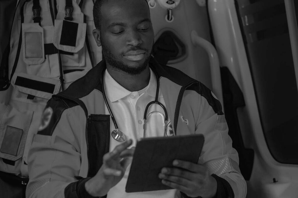 A person in a medical uniform with a stethoscope around their neck uses a tablet inside an ambulance.
