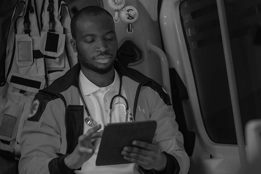 A medical professional in a uniform and stethoscope is seated inside an ambulance, using a tablet.