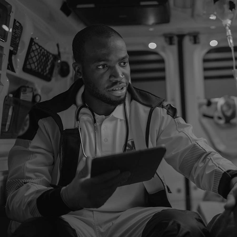 Paramedic in uniform sits inside an ambulance, holding a tablet and looking to the side.