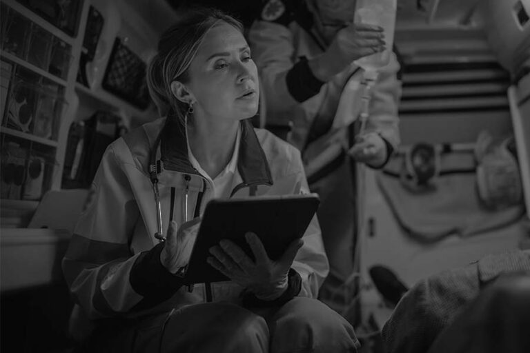 Paramedic in an ambulance holds a tablet, writing notes. Another paramedic in the background adjusts an IV drip near a patient.