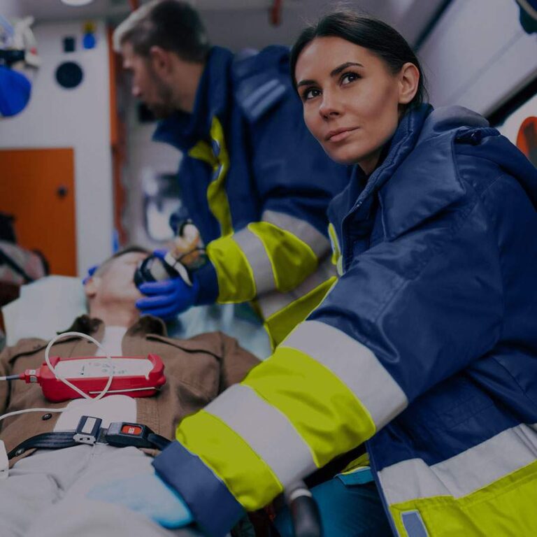 Paramedics in blue and yellow jackets attend to a patient on a stretcher inside an ambulance, with medical equipment visible.