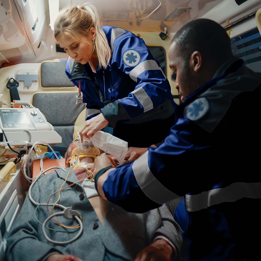 Two paramedics in an ambulance provide medical care to a patient lying on a stretcher. Equipment and medical devices are visible around them.