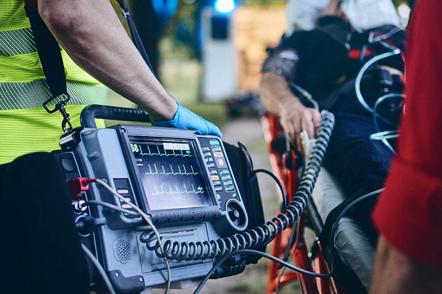 A paramedic monitors a patient's vital signs on a medical device while the patient lies on a stretcher outdoors.