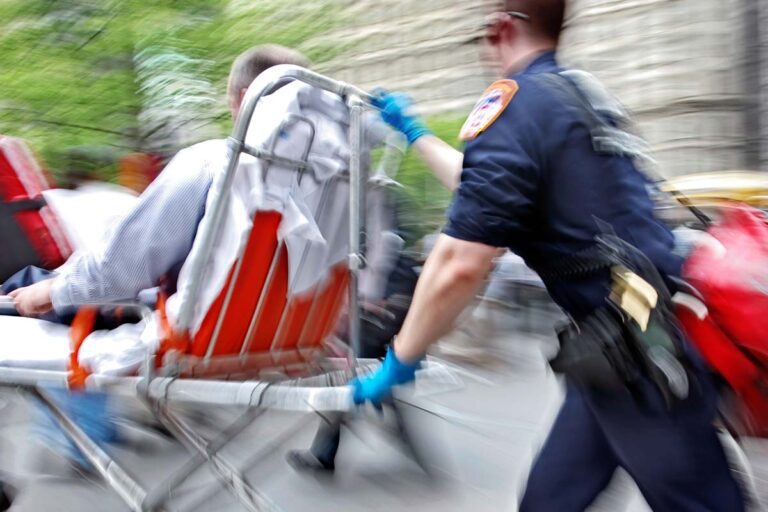 Emergency responder rushes a person on a stretcher down a city street, with both moving quickly past blurred surroundings.