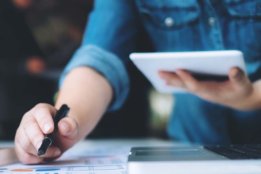A person in a denim shirt writes with a pen and holds a tablet, with papers and a laptop nearby.