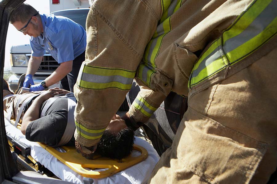 Emergency responders assist a person on a stretcher next to a vehicle. One responder is adjusting a neck brace while another tends to the patient's side.