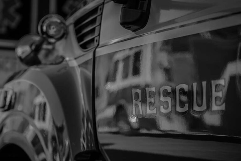 Close-up of a rescue vehicle with the word "RESCUE" visible on its side, featuring a chrome headlight and side mirror. Image is in black and white.
