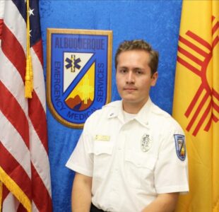 A person in an emergency medical uniform stands between the American flag and the New Mexico state flag, with an Albuquerque Emergency Medical Services plaque in the background.