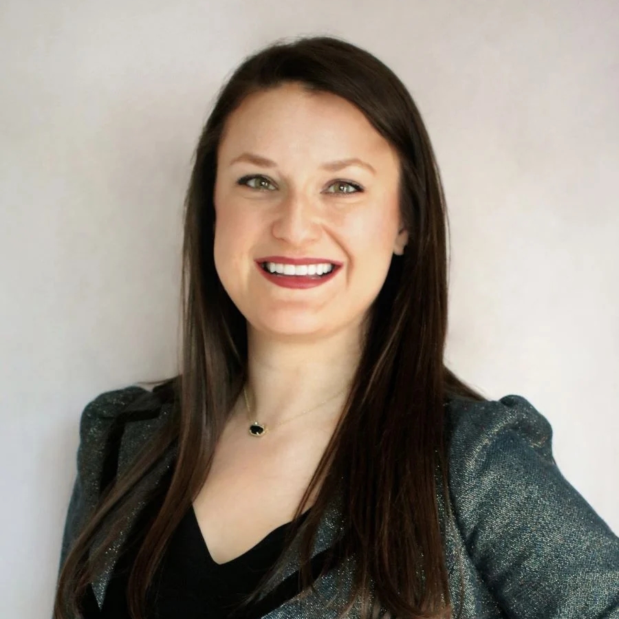 Alyssa M. Green, a smiling person with long brown hair, wearing a dark top and a shimmering jacket, against a light background.