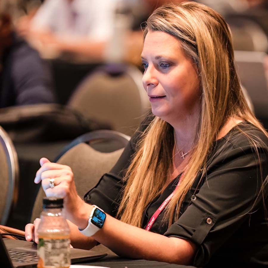 A person with long hair wearing a smartwatch is sitting at a table, engaged with a laptop. A water bottle is on the table.