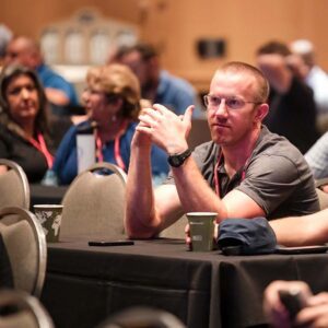 A man with glasses sits at a table in a conference room, watching attentively. People in the background are seated and engaged in conversation.