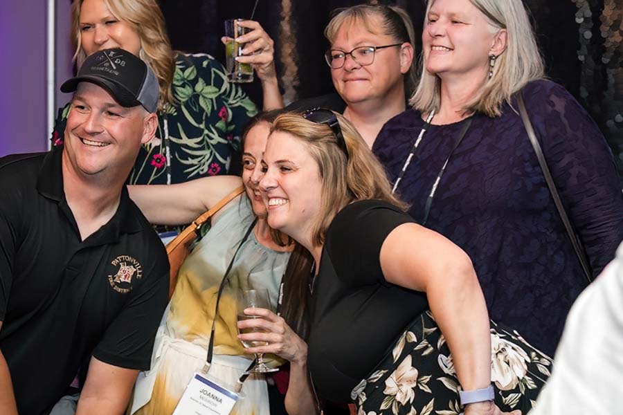 Group of people smiling and posing together at an indoor event. Some hold drinks and wear event badges.