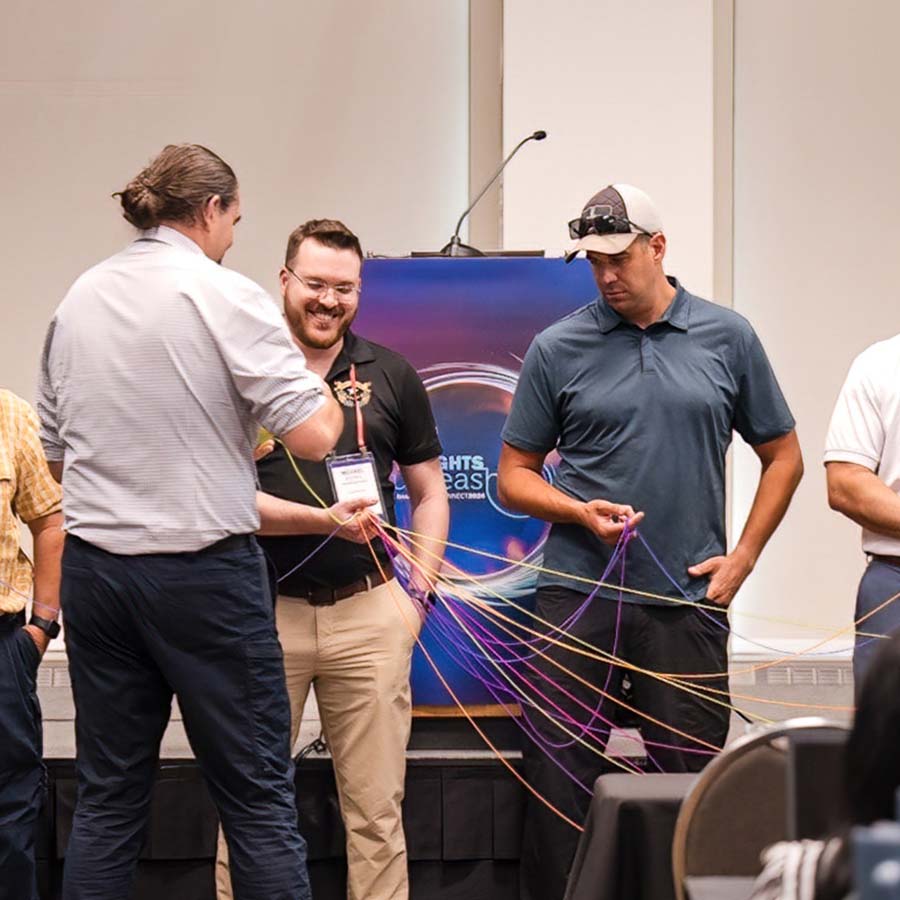 Men standing in a room, interacting with colorful strings. A podium with a logo is in the background.