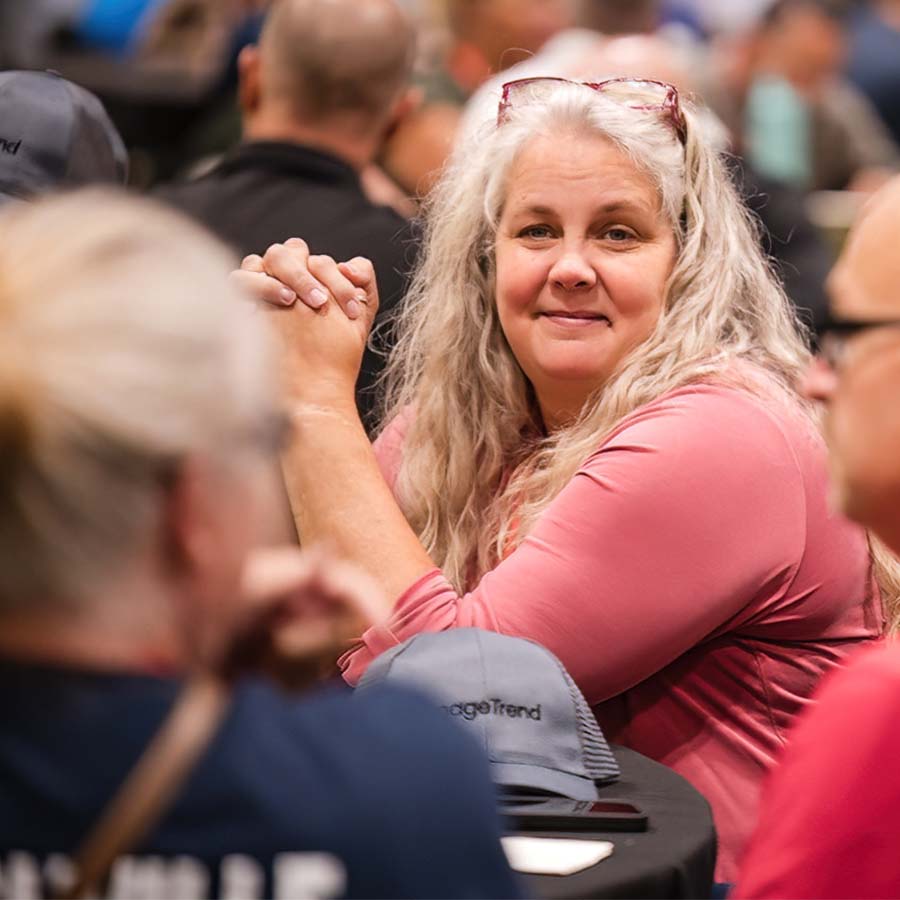 A woman with long hair sits at a table, resting her hands on it, in a crowded indoor setting.