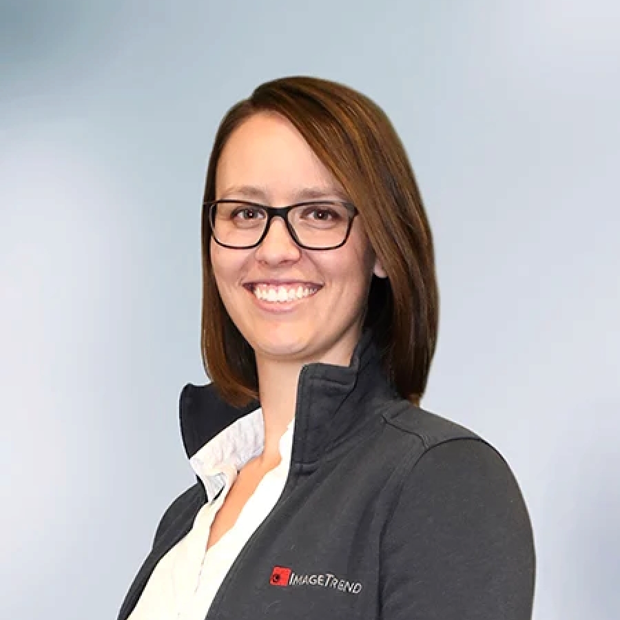 Morgan K. Anderson, a person with shoulder-length brown hair and glasses smiles, wearing a dark jacket over a white shirt, standing against a plain background.