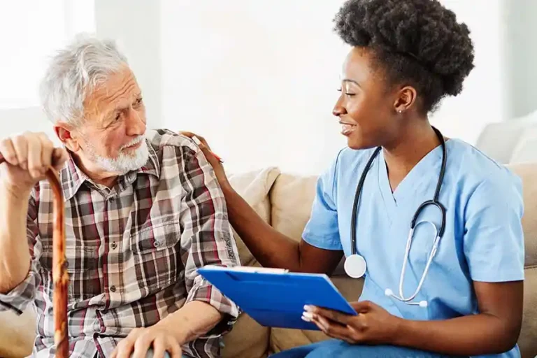 A nurse with a stethoscope talks to an elderly man holding a cane, while holding a clipboard.