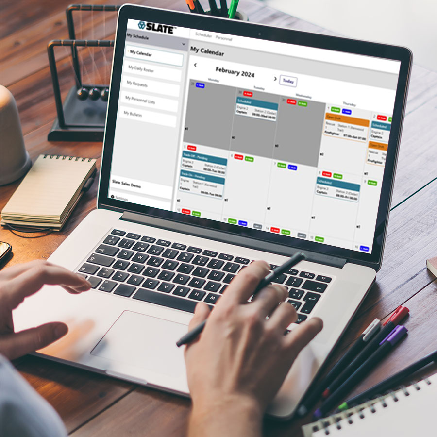 Person using a laptop with a digital calendar open for February 2024. The screen shows multiple color-coded events. The desk has notebooks and a pen.