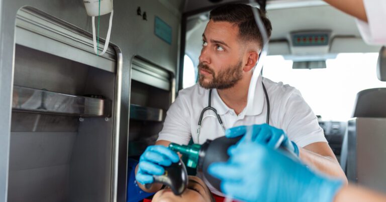 Paramedic in an ambulance, wearing a stethoscope, operates a manual resuscitator. Blue-gloved hands assist nearby.
