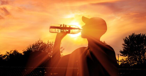 A person in a cap drinks from a bottle with the sun setting behind them, creating a silhouette and sunray effect.