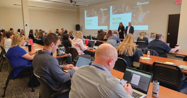 Conference room with attendees seated, some using laptops. Two presenters stand at the front near a screen displaying "Welcome Audience.