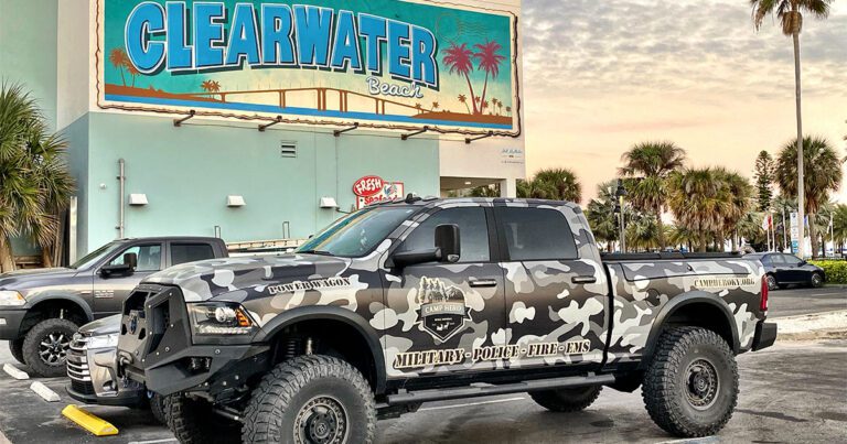 Camouflage-patterned pickup truck parked near a large "Clearwater Beach" sign with palm trees and a sunset in the background.