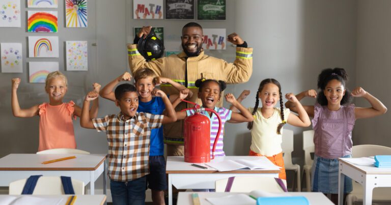 A firefighter stands with smiling children flexing their arms in a classroom. Artworks and letters are on the wall behind them, and there are desks and chairs in the foreground.