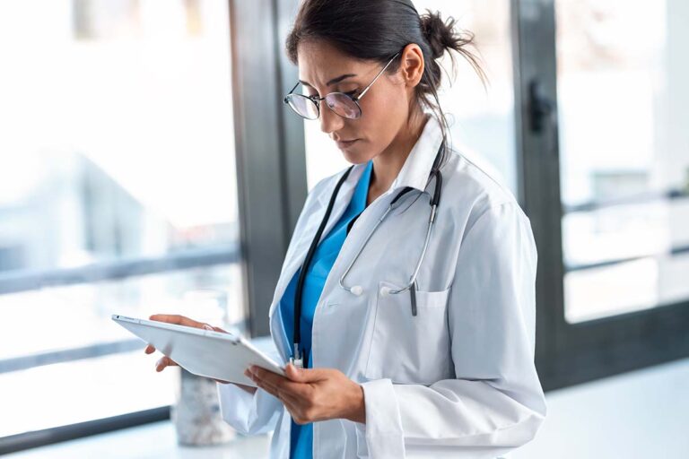 A woman wearing glasses, a white coat, and a stethoscope stands in a well-lit room, looking at a tablet.
