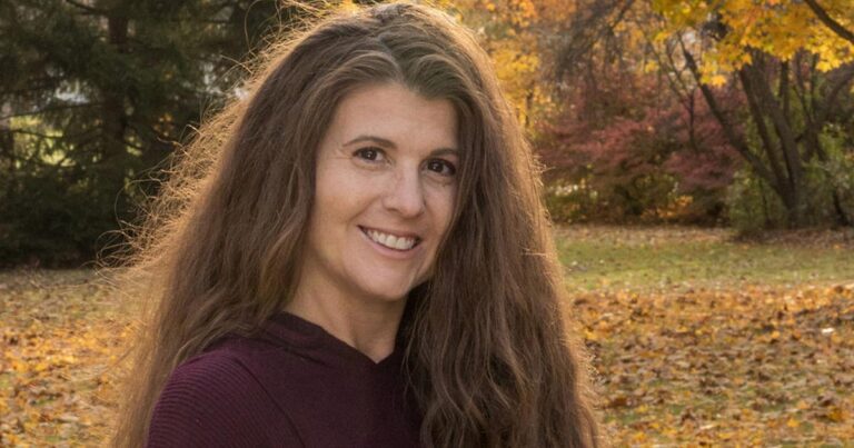A woman with long brown hair smiles while standing in a park with autumn foliage in the background.