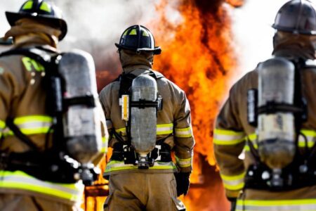 Three firefighters in gear stand facing a large fire.