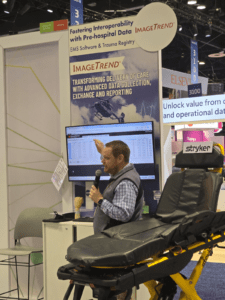 A man with a microphone speaks in front of a screen displaying data, standing beside a stretcher at an expo booth for ImageTrend, which focuses on EMS software and trauma data solutions.