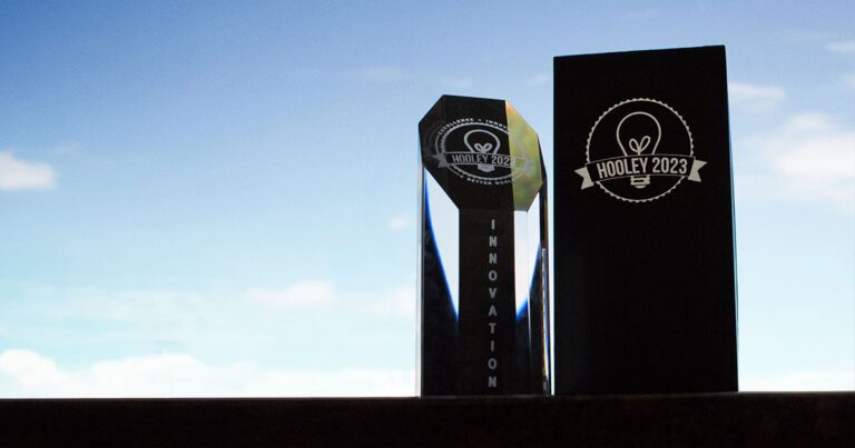 A glass trophy labeled "Hooley 2023 Innovation" stands next to a tall black box against a blue sky background.