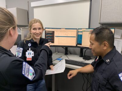 Three EMTs in uniform are discussing something on computer screens. One is holding a document, and they appear to be smiling and engaged in conversation in an office setting.