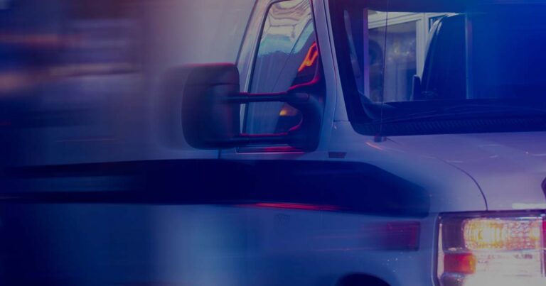Close-up of an ambulance with emergency lights on. The side mirror and part of the front cab are visible. Blurred lights create a sense of motion.