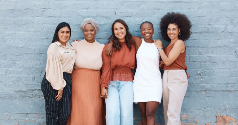 Five women standing together in front of a blue brick wall, smiling and casually dressed in neutral tones.