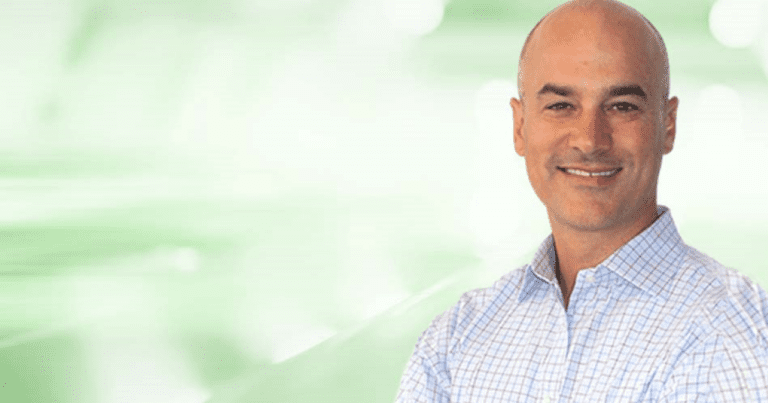 Bald man smiling, wearing a checkered shirt against a blurred green background.