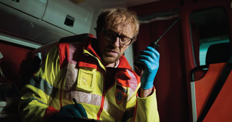 Paramedic wearing high-visibility jacket and blue gloves, holding a walkie-talkie, and writing on a clipboard inside an emergency vehicle.