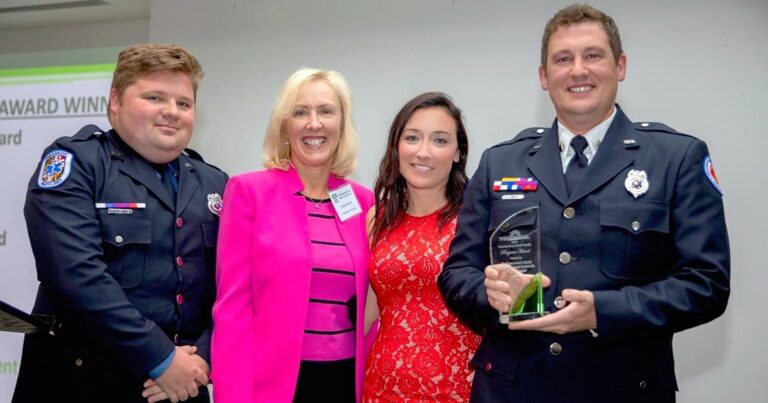 Four people stand together, two in uniforms and two in civilian clothing. One uniformed person holds an award. They are posing for a photo indoors.