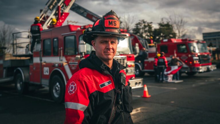 A firefighter in a red and black uniform stands in front of red fire trucks with extended ladders, outdoor setting, cloudy sky.