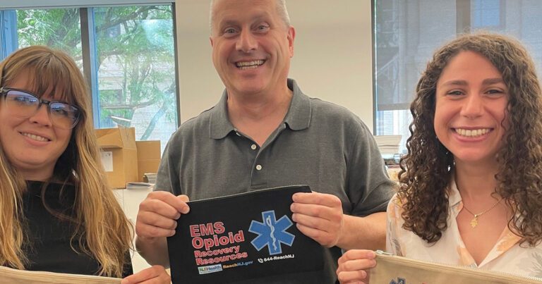 Three people smiling and holding pouches related to EMS opioid recovery resources in an office setting.