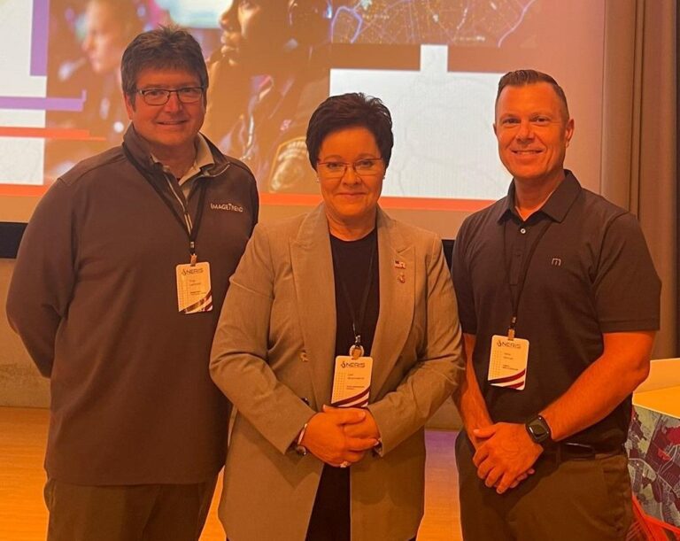 Three people in lanyards stand together in a conference setting with a presentation screen behind them.