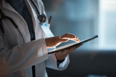 A person wearing a white lab coat and stethoscope uses a tablet, illuminated by its screen in a dim setting.