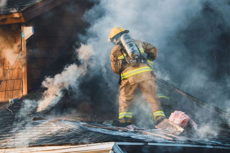 A firefighter in gear hoses down a smoking rooftop, surrounded by heavy smoke, during daylight.