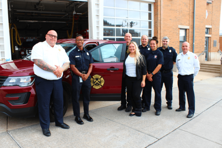 A group of eight people, including firefighters in uniform, stand in front of a parked fire department SUV near a building.