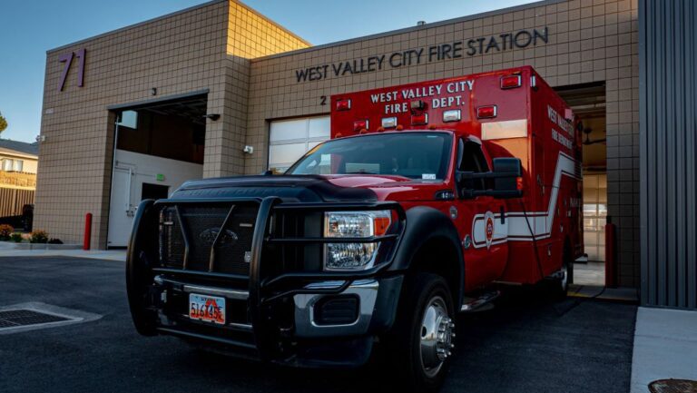 Red fire department vehicle parked outside West Valley City Fire Station with open garage doors.