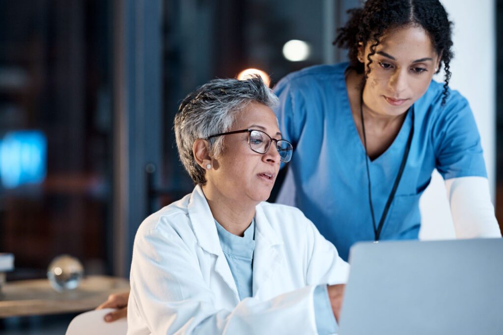 Two healthcare professionals in blue and white attire look at a laptop screen intently, analyzing market intelligence data to enhance patient care.