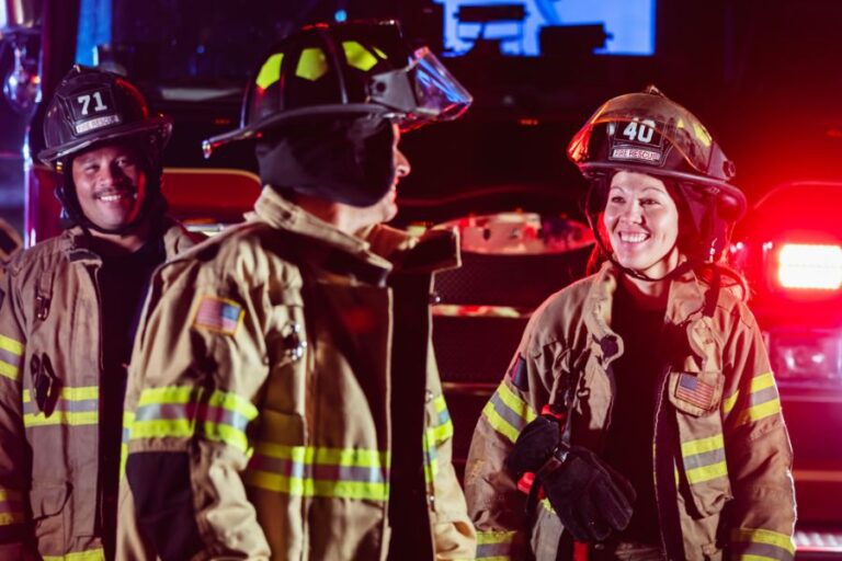 Three firefighters in gear stand in front of a fire truck at night, smiling and interacting.