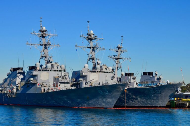 Two large naval warships docked side by side at a harbor on a clear day, with multiple antennas and radars visible on their decks.